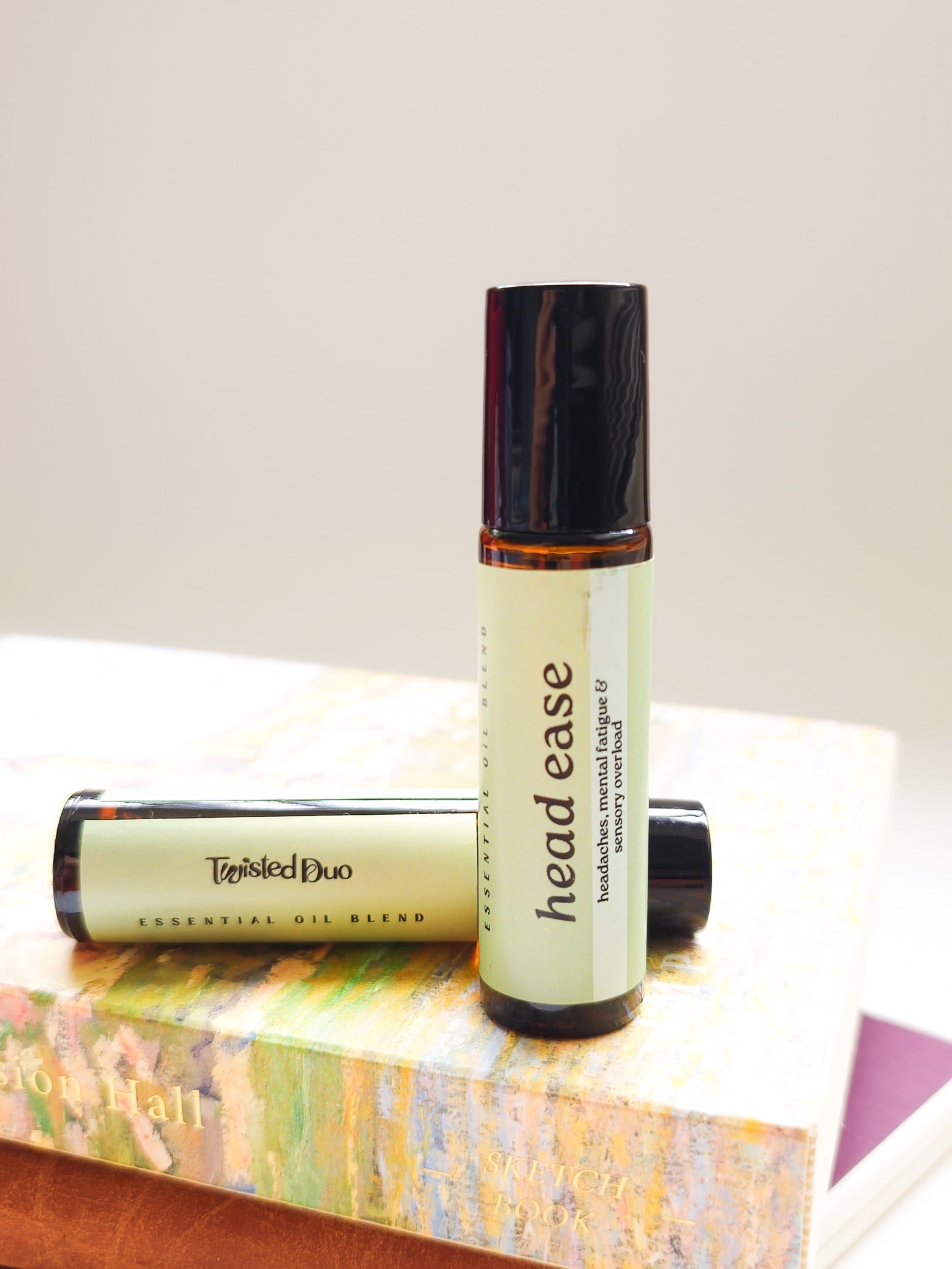 Two bottles of 'head ease' essential oil blend on a book with a white background