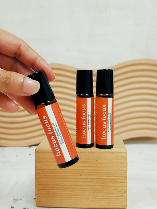 Hand holding a 'hocus focus' rollerball next to two other similar products on a wooden stand.