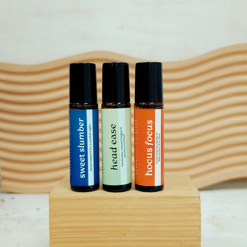 Three roller bottles labeled 'sweet slumber', 'head ease', and 'hocus focus' on a wooden stand with a wavy wooden background.