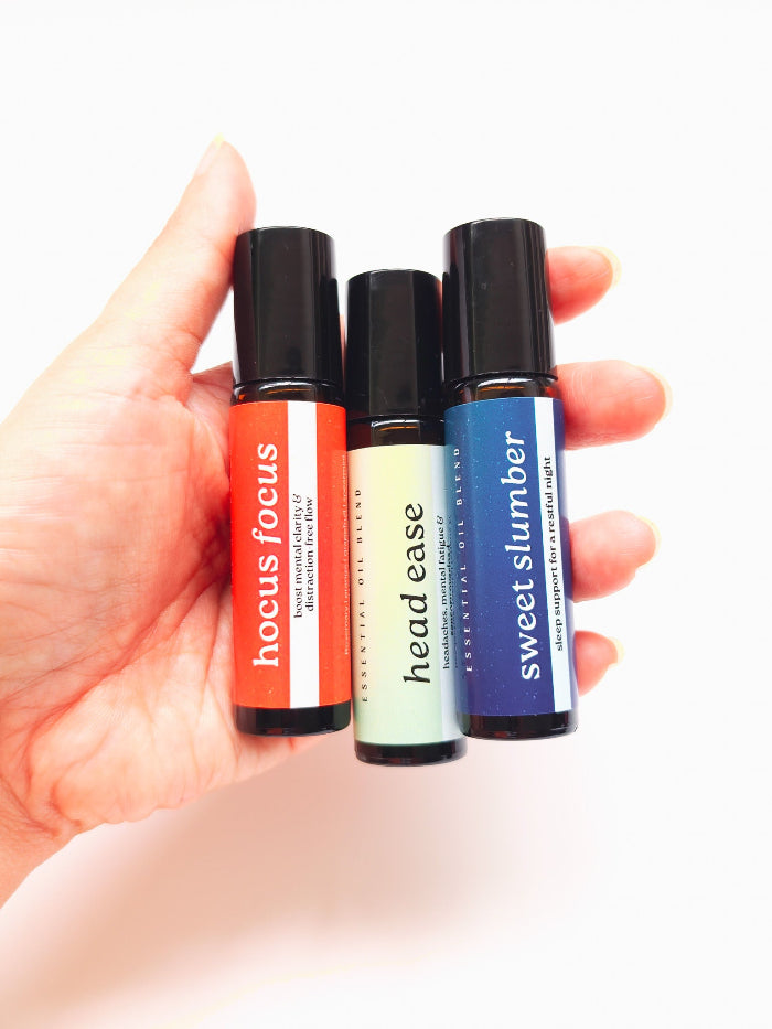 Three small rollerball bottles labeled 'hocus focus', 'head ease', and 'sweet slumber' held in a hand against a white background.